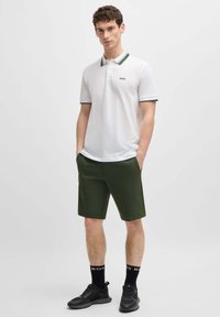 White polo shirt with green striped collar, paired with dark green shorts. Model stands with hands in pockets, wearing black sneakers and socks.