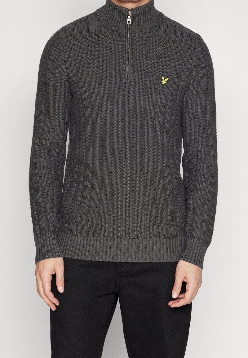 Gestrickter grauer Pullover mit hohem Kragen und Viertelreißverschluss, der eine gerippte Textur und ein kleines gelbes Logo auf der Brust aufweist.