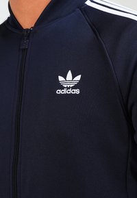 Giacca da allenamento Adidas blu navy con colletto alto, chiusura con zip e tre strisce bianche sulle spalle, con logo bianco sul petto.