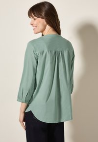 Blusa verde chiaro con scollo rotondo, maniche a tre quarti, dettagli plissè sulla schiena e orlo arrotondato. Il tessuto appare liscio e leggero.