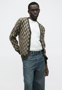 Cardigan à carreaux marron et beige à coupe décontractée, en tissu doux. Porté sur un t-shirt blanc, associé à un jean bleu. Boutons visibles.