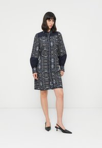 Woman wearing a knee-length navy floral dress with long puffed sleeves and black pointed slingback heels standing against a white background.