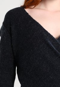 Pull en maille noire avec un décolleté en V profond, présentant une texture douce et des manches longues, affichant une finition légèrement duveteuse.