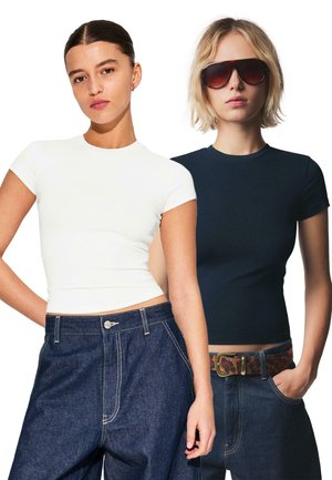 Weiße und marineblaue figurbetonte T-Shirts mit kurzen Ärmeln; getragen zu hoch taillierten Jeans. Ein Model hat blonde Haare und trägt eine Sonnenbrille.