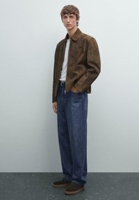Giacca in suede marrone, T-shirt bianca, jeans larghi blu e scarpe marroni scure. Modello in piedi contro uno sfondo grigio chiaro.