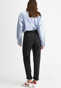 Persona che indossa una camicia a righe blu con maniche lunghe, jeans neri a vita alta con i risvolti, e sneakers bianche con dettagli rossi, in piedi di spalle.