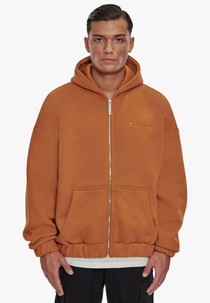 Lynlukkede hettejakke i brun-oransje stoff, med en frontlomme, ribbestrikkede mansjetter og et brodert logo på venstre bryst.