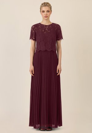 Frau mit blondem Haar trägt ein bordeauxrotes Spitzenkleid mit kurzen Ärmeln und einem plissierten langen Rock, steht vor einem beigen Hintergrund.