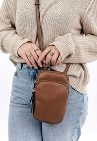 Borsa a tracolla in pelle marrone con una finitura testurizzata, forma rettangolare e dettagli in metallo argentato sulla cerniera. Indossata con un maglione a maglia e jeans.