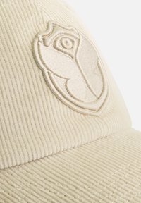 Casquette en velours côtelé crème avec un emblème brodé d'une rose stylisée à l'intérieur d'un blason. Texture côtelée distinctive sur la surface.