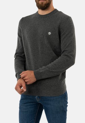 Grauer Strickpullover mit rundem Halsausschnitt und gerippten Bündchen, ausgestattet mit einem kleinen Logo auf der linken Brust, kombiniert mit blauen Jeans.