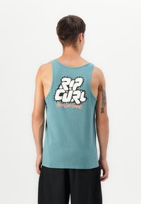 Muškarac s kratkom smeškom kosom koji nosi tirkiznu majicu bez rukava s natpisom "Rip Curl Hardcore Surfing" na leđima i crne šortse, na bijeloj pozadini.