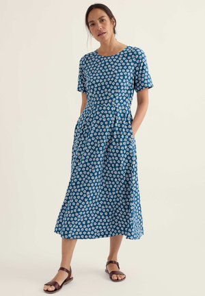 Žena v modrých midi šatách s bielym motívom margarétok, krátkymi rukávmi a vreckami, v hnedých sandáloch na plochom podpätku, stojaca.