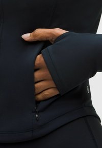 Fekete cipzáras sportdzseki, amely illeszkedő fazonú, texturált anyagból készült, és rejtett zseb található a deréknál, sima cipzárral záródik.