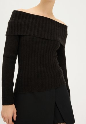 Femme portant un pull noir côtelé épaules dénudées à manches longues et une jupe noire sur un fond clair uni.