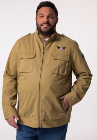 Giacca beige con chiusura a zip frontale, due tasche sul petto e tasche laterali. Presenta un logo alato sul lato sinistro del petto e una toppa rotonda sulla manica.