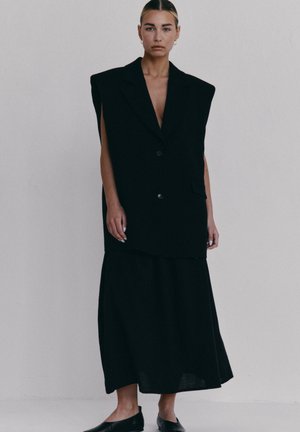 Kobieta stojąca na tle gładkiej ściany, ubrana w oversize'owy, bezrękawnikowy czarny żakiet oraz długą czarną spódnicę, w czarnych butach wsuwanych.