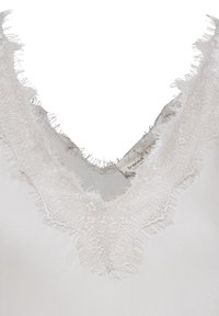 Vêtement blanc à encolure en V avec une délicate bordure en dentelle effilochée autour du col et une petite étiquette de marque visible à l'intérieur du col.