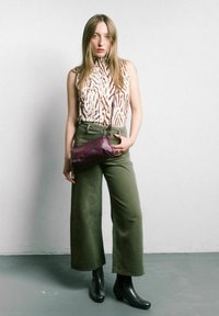 Top senza maniche color beige chiaro con motivo a zebre marrone, pantaloni larghi color verde oliva e stivali neri alla caviglia. Una borsa a tracolla viola aggiunge un tocco di colore.