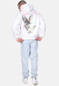 Un hoodie blanc avec un graphique d'aigle et le texte "Saison de chasse" en rose, associé à un jean bleu clair et des baskets blanches.