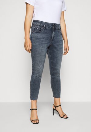 Eine Frau trägt dunkelgraue Skinny-Jeans mit ausgefransten Säumen, ein weißes Kurzarmshirt und schwarze High-Heel-Sandalen und steht vor einem schlichten Hintergrund.
