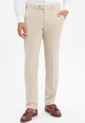 Beige, maßgeschneiderte Hose mit Vorderfalten, getragen mit braunen Quasten-Loafers und einem weißen Hemd, eine Hand in der Tasche.