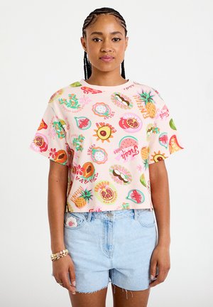 Vrouw met gevlochten haar die een pastelroze T-shirt draagt met diverse kleurrijke fruitdessins en lichtblauwe denimshorts.