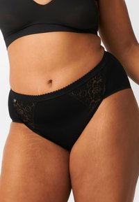 Mutande nere con design a vita alta, dotate di un inserto in pizzo sui lati. Tessuto liscio con bordi scallopati lungo la cintura.