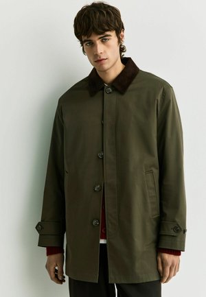 Young man with brown wavy hair wearing an olive green buttoned coat with dark collar and red sweater sleeves visible.