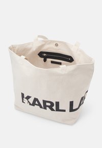 Beżowa torba materiałowa z czarnym napisem "KARL LAGERFELD", otwarta u góry, z kieszenią na zamek błyskawiczny wewnątrz oraz zapięciem na zatrzask.