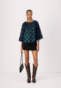 Haut texturé bleu marine et sarcelle avec un motif à bords festonnés, manches larges, associé à une jupe noire et des bottes de cowboy marron, tenant un petit sac à main.