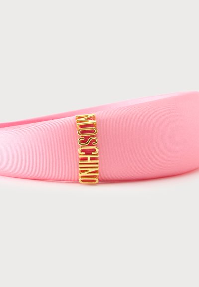 Bandeau en tissu rose lisse avec des lettres métalliques dorées verticales formant le mot "MOSCHINO", attaché d’un côté.