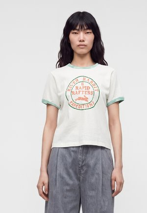 Jeune femme aux longs cheveux foncés porte un t-shirt blanc avec une bordure verte et un texte orange indiquant "River Rabbit & Rapid Rafters Expeditions."