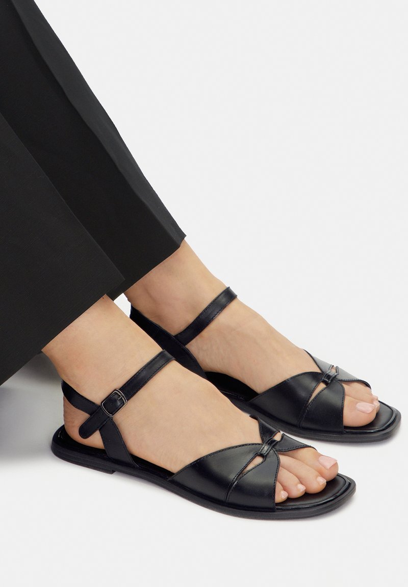 Sandalias de cuero negro con diseño de tiras cruzadas, dedos abiertos, correas en el tobillo y suela plana. Textura suave con acentos mínimos de herrajes.