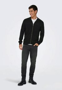 Juodas užtrauktuku užsegamas megztinis, pagamintas iš megzto audinio, turintis prigludusį siluetą, gumuotus rankogalius ir glotnią tekstūrą. Dėvimas su pilkais džinsais ir juodais batais.