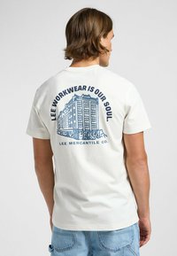 En creamfarvet bomulds T-shirt med et blåt grafik af en bygning og tekst på bagsiden. Kortærmet og med standart pasform.