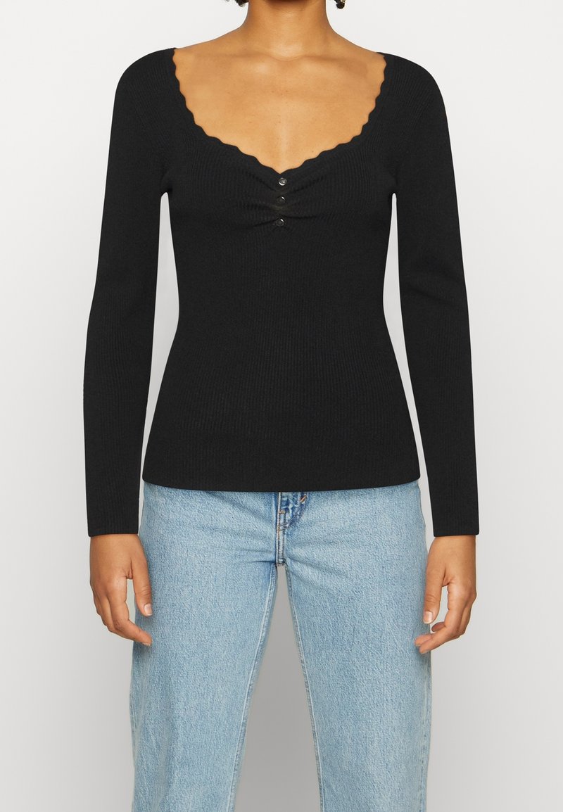 Schwarzes, geripptes Langarmshirt mit gewelltem Ausschnitt und drei vorderen Knöpfen, kombiniert mit hellblauen Jeans.