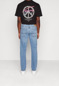 Černé tričko s krátkým rukávem a symbolem míru, s grafickým textem na zadní straně, v kombinaci s světle modrými rovnými džíny a bílými botami.