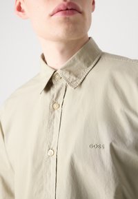 Camisa de botonada color beige claro con una textura suave, que presenta un collar clásico y un pequeño logo verde de "BOSS" en el pecho.