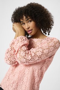 Top en dentelle rose avec un motif floral, des manches longues bouffantes et un col rond. Le tissu a une apparence texturée et aérée.
