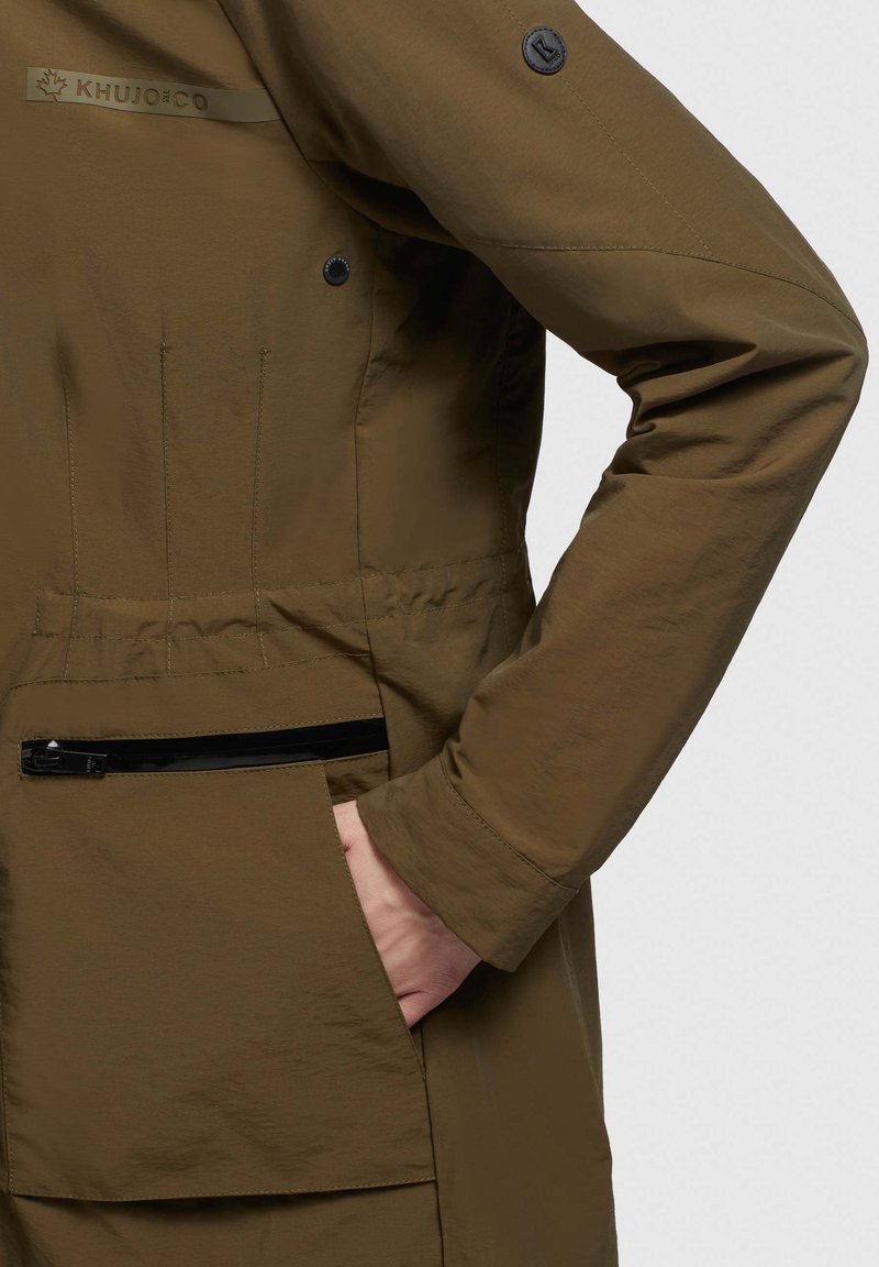 Khujo-Jacke in Olivgrün, aus leichtem Stoff gefertigt. Verfügt über eine Seitentasche mit einem schwarzen Reißverschluss und eine Hand, die in der unteren Tasche ruht.