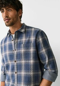Kék kockás ing texturált anyagból, elöl zsebbel. Gombos gallérral és kontrasztos kockás mintával a mandzsettán.