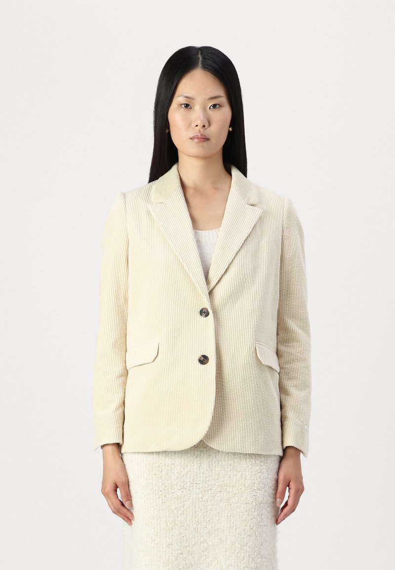 Beige corduroy-takki, jossa on viistot kaulukset, kaksi etutaskua ja kaksinappinen kiinnitys. Rakenne, jossa on pystysuorat urat.
