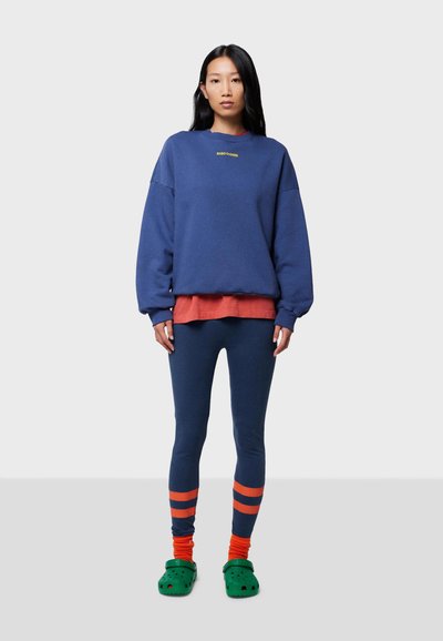Blå oversized sweatshirt, röd lagerblus, marinfärgade leggings, orange- och marinstregrade strumpor, gröna träsulor, mjuka tygtexturer.