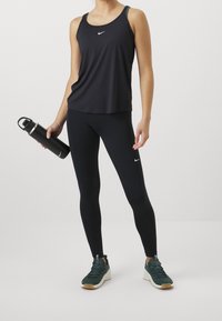 Débardeur de sport noir avec logo Nike, associé à un legging noir. Le modèle tient une bouteille d'eau noire. Baskets vertes avec semelle texturée.