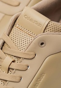 Zapatilla beige de Calvin Klein con parte superior texturizada, perforaciones, cordones de textil y un logo en relieve en la pestaña del talón.