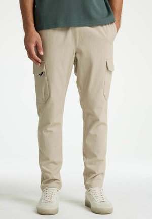 Muž v béžových nákladných nohaviciach s prekrytými vreckami a bielych teniskách, stojaci s voľne spustenými rukami pozdĺž tela.