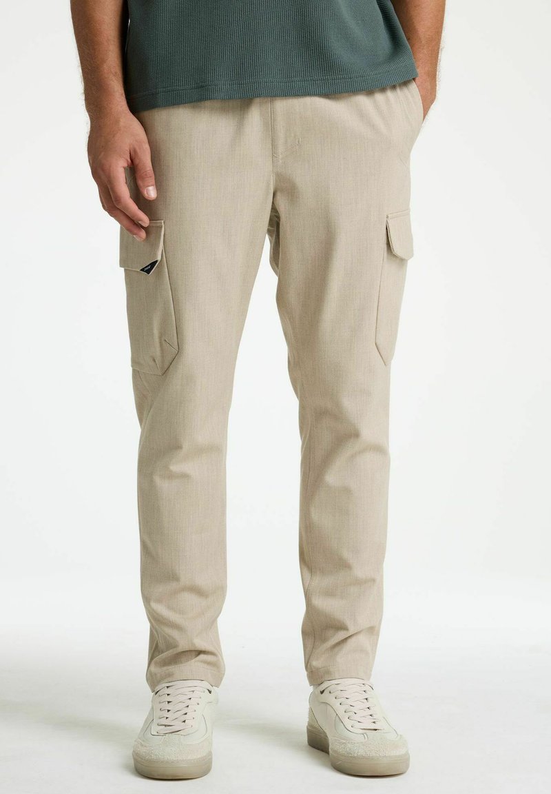 Mees, kes kannab beeže lahtiste taskutega tööpükse ja valgeid tossusid, seisab käed lõdvalt kõrval.