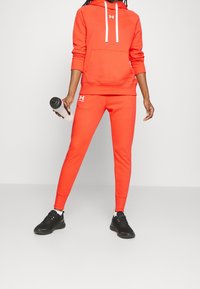 Felpa sportiva rossa con cappuccio e lacci bianchi, abbinata a jogger fitted dello stesso colore. Sneakers nere e una bottiglia d'acqua completano l'outfit.