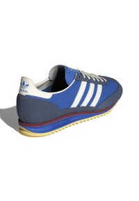 Blue sneaker with gray accents, three white stripes, textured blue toe, and yellow sole. Features a retro athletic design and classic branding.